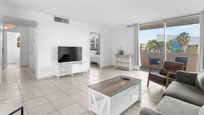 A modern living room with a flat-screen TV, a coffee table, and a sofa. There is a balcony with a view of palm trees and buildings. - Bay Harbor Sunset Apt 3A (Bay Harbor Islands)