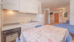 A compact kitchen with white cabinets, a built-in oven, and a dining area with a table covered in a patterned tablecloth. - APPARTAMENTI GLI ORIZZONTI - ORIZZONTI N MONO 15 (Marina di Campo)