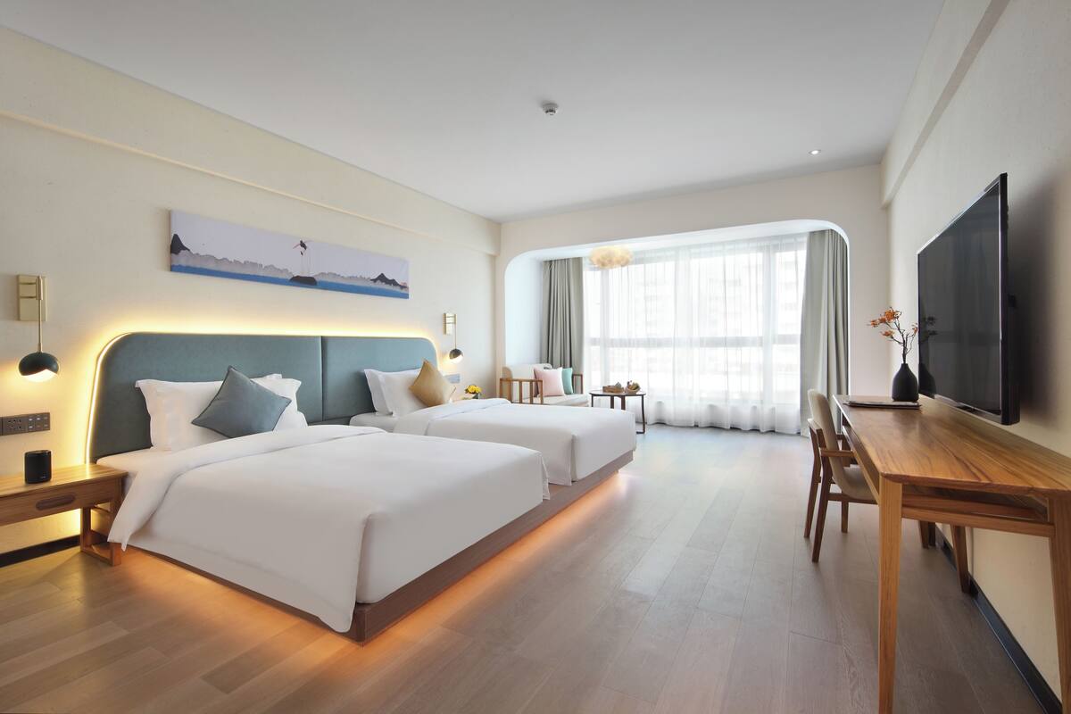 A hotel room with two beds, a desk, a TV, and a large window with curtains.