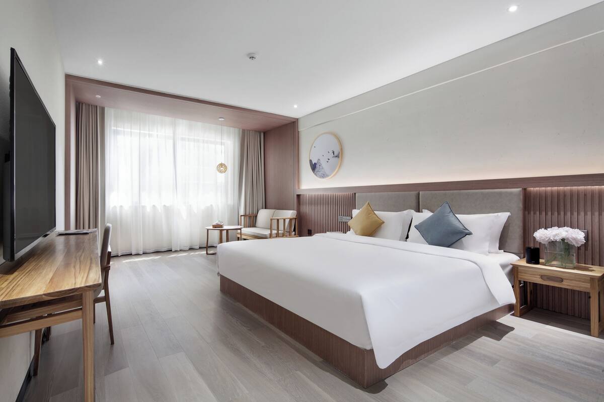 A modern hotel room with a large bed, a desk, a TV, and a window with curtains.