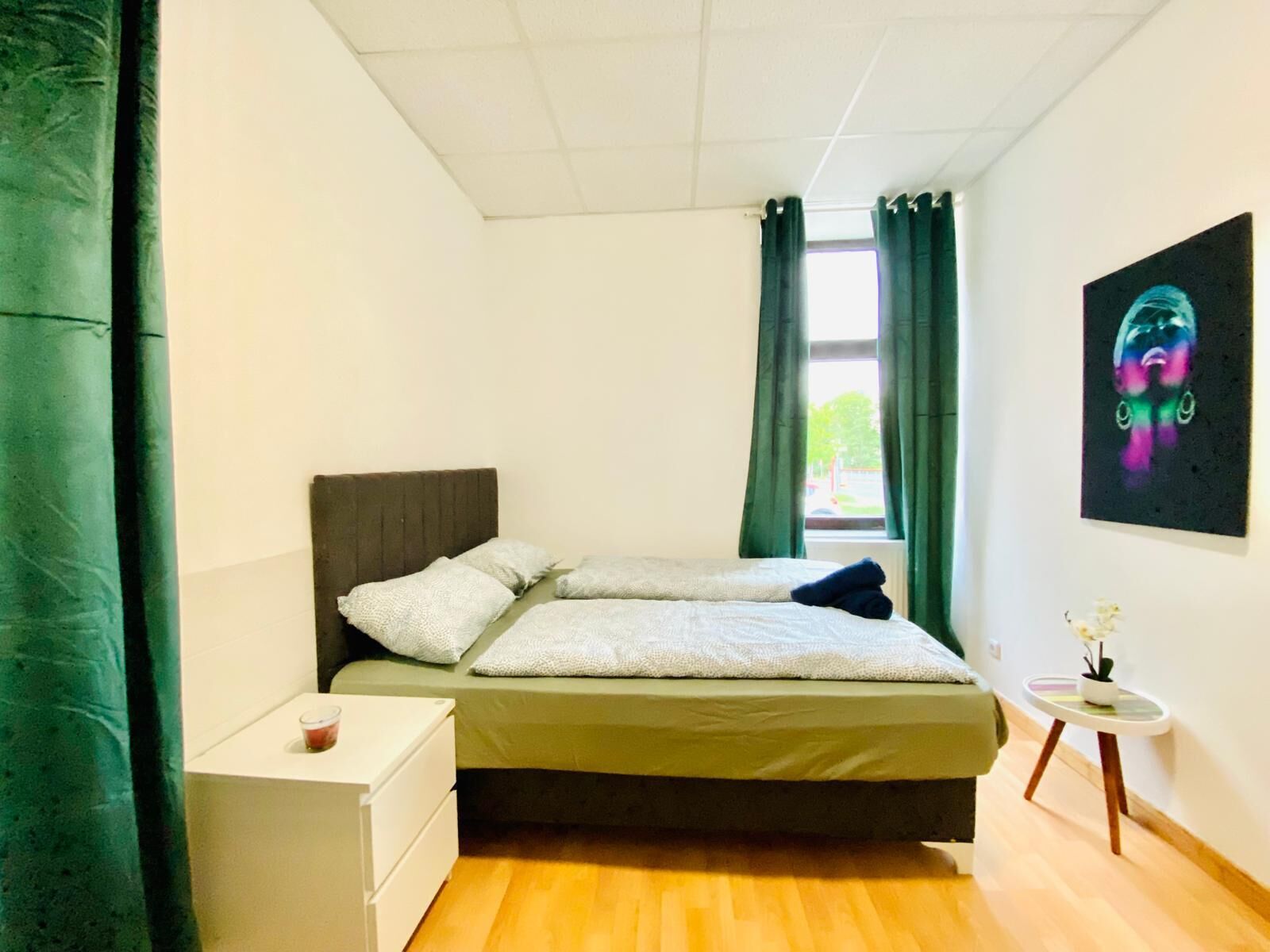 A small, neatly arranged bedroom with a bed, a bedside table, a small table with a plant, and a window with curtains.