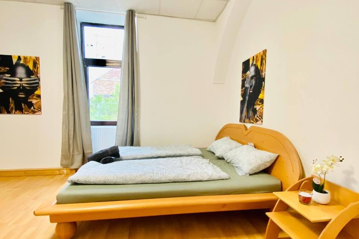 Ein modernes Schlafzimmer mit Holzboden, einem Bett mit grüner Matratze und weißen Kissen, einem Nachttisch aus Holz mit einer kleinen Tischplatte und einer Blumenvase sowie zwei abstrakten Gemälden an den Wänden.
