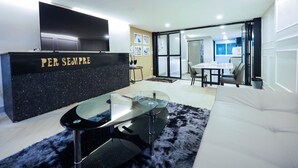 Een moderne woonkamer met een glazen salontafel, een zwarte bank en een flatscreen televisie aan de muur.