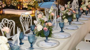 Una larga mesa de comedor dispuesta para un evento formal, con sillas blancas y cristalería azul, decorada con flores y velas, con vistas a un paisaje pintoresco.