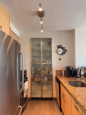 Une cuisine compacte équipée d’un réfrigérateur en acier inoxydable, d’un meuble vitré à deux portes, d’une horloge murale et d’une machine à café posée sur le plan de travail.