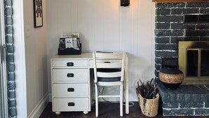 Een kamer met een witte wand, een zwart metselwerk haard, een wit bureau met lades, een witte stoel, een wandlamp en een televisie aan de muur.