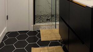Een badkamer met een douchecabine van glas, een witte deur, een zwart-wit betegelde vloer en een zwart wastafelmeubel met een wit aanrecht.