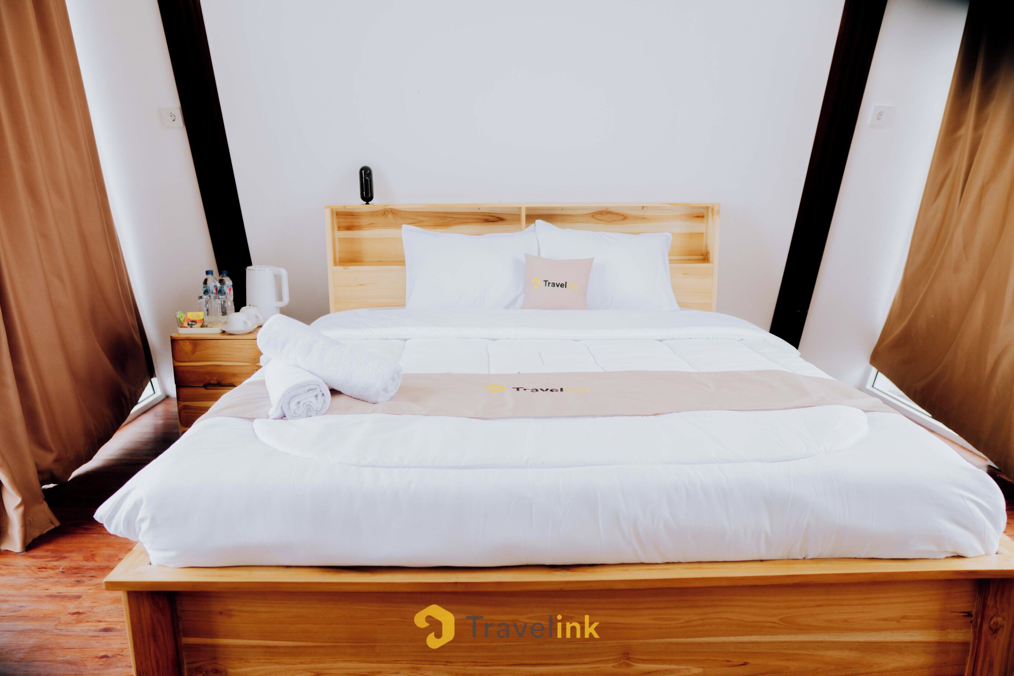 A neatly made bed with white linens, a wooden headboard, and a Travelink logo on the bedspread.
