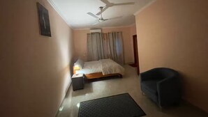 Une chambre d’hôtel avec un lit, un bureau avec une lampe, une chaise, un ventilateur de plafond et une fenêtre avec des rideaux.