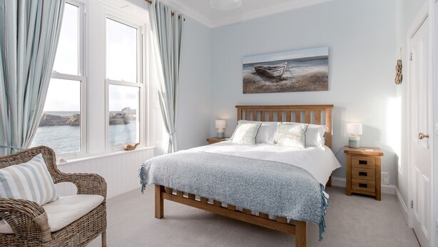 A bedroom with a large bed, a wicker chair, a nightstand, a painting of a boat on the wall, and a view of the sea through the window. - Portpatrick House by Interhome (Portpatrick)