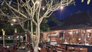 An outdoor dining area with wooden tables and chairs, a tree, and a building with a thatched roof in the background. - Balkondes Tuksongo (Borobudur)