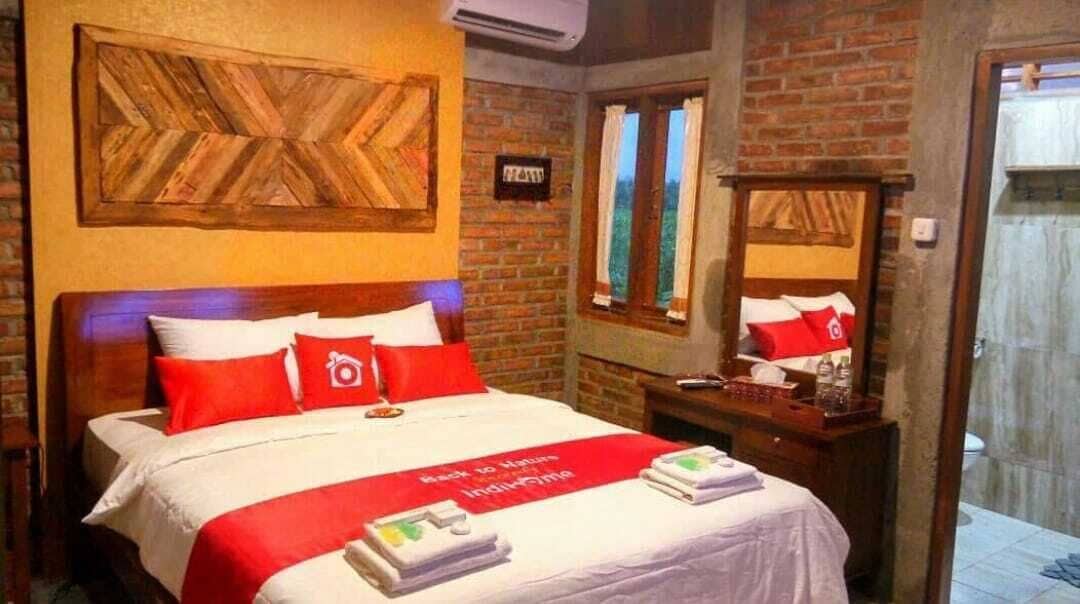 A hotel room with a brick wall, a large bed with red and white bedding, a wooden headboard, a bedside table with a lamp, and a window with curtains.
