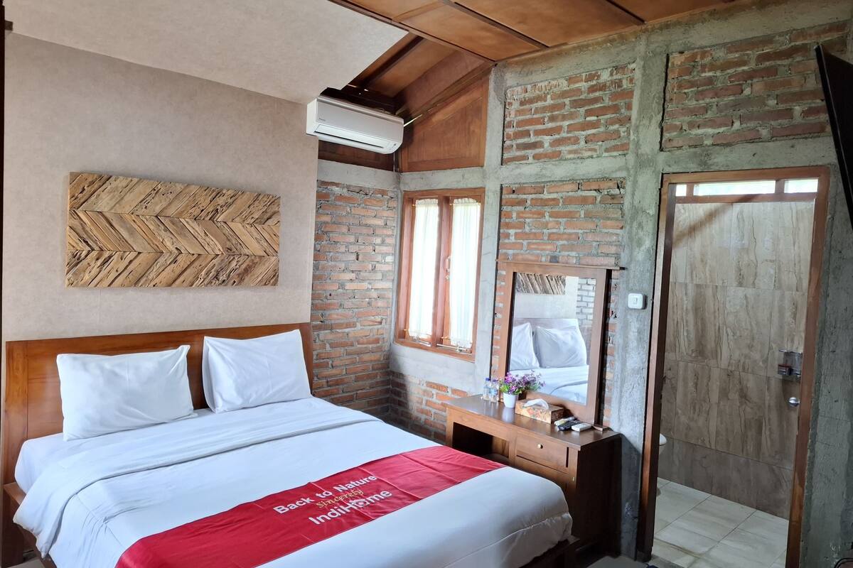 A bedroom with a bed, a wooden headboard, a brick wall, a wooden desk, a mirror, and a door.