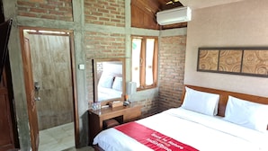 A hotel room with a brick wall, wooden ceiling, a bed with white bedding and a red throw, a wooden desk, a mirror, and a window. - Balkondes Tuksongo (Borobudur)