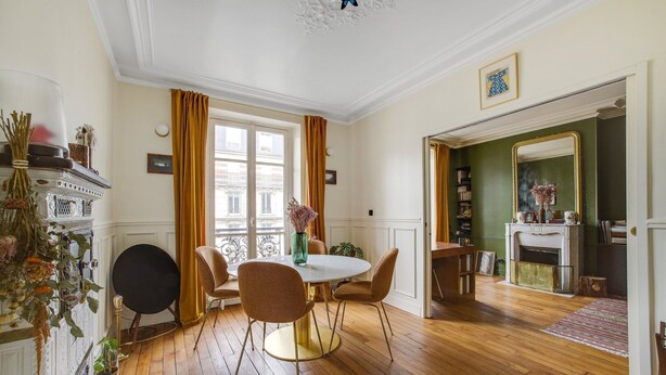 Ein Essbereich mit einem runden Tisch, Stühlen und einem Blumenstrauß. Es gibt ein Fenster mit Vorhängen, einen Kamin und ein Bücherregal.