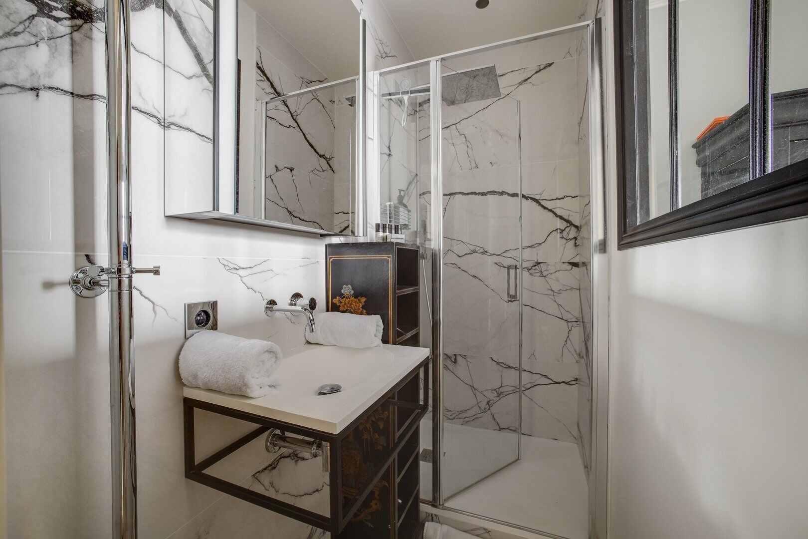 A bathroom with a marble-tiled shower enclosure, a white sink with a mirror, and a small cabinet.