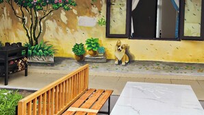 A cozy outdoor seating area with a wooden bench and a table, a potted plant, and a dog sitting by a wall with a painted tree and flowers. - Mit homestay (Hue)