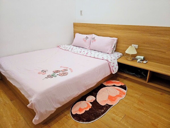 A bedroom with a wooden headboard, a bed with pink bedding, a bedside lamp, and a small shelf with a lamp and a phone.
