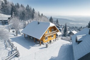Una cabaña de madera con techo nevado y un balcón, rodeada de un paisaje invernal y árboles.