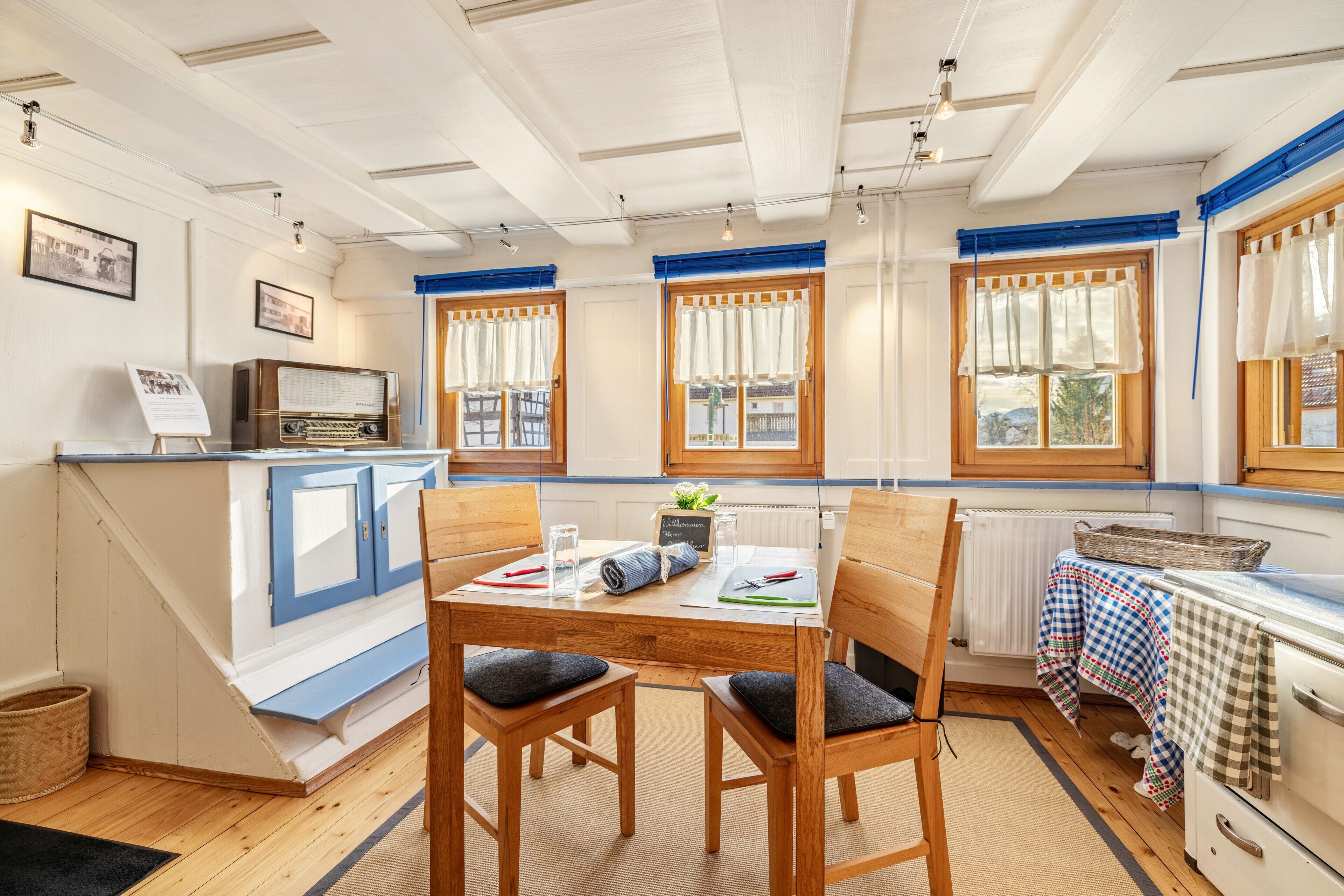A dining area with a wooden table, chairs, a radio, and a view of the outside through windows.