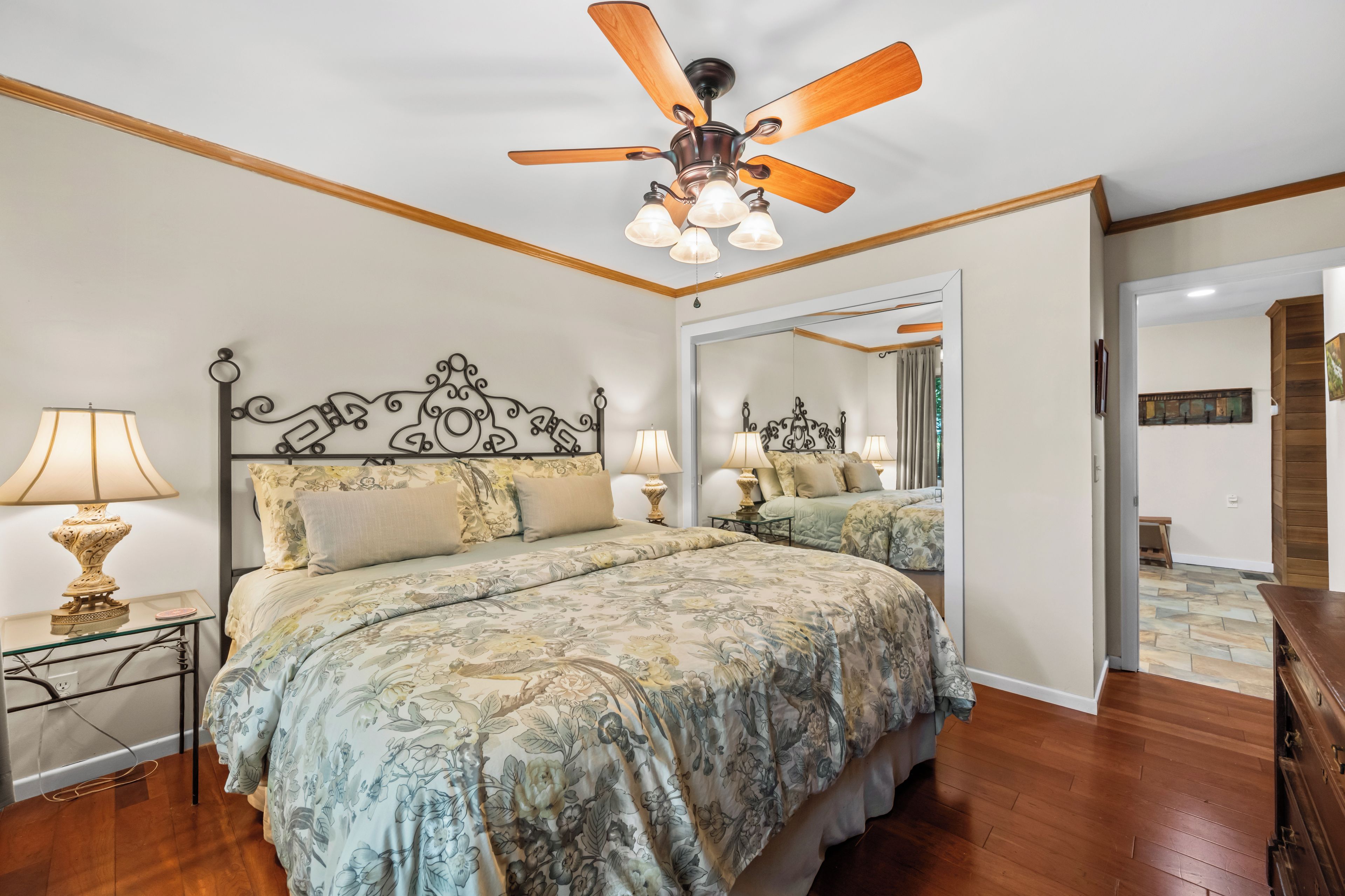 A bedroom with a bed, a ceiling fan, a nightstand with a lamp, and a mirror.