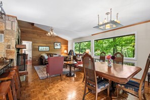 A spacious living area with a wooden floor, a stone wall, a fireplace, and a large window offering a view of greenery.