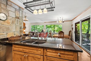 A kitchen with a stone wall, wooden cabinets, a double sink, and a large window overlooking a deck.