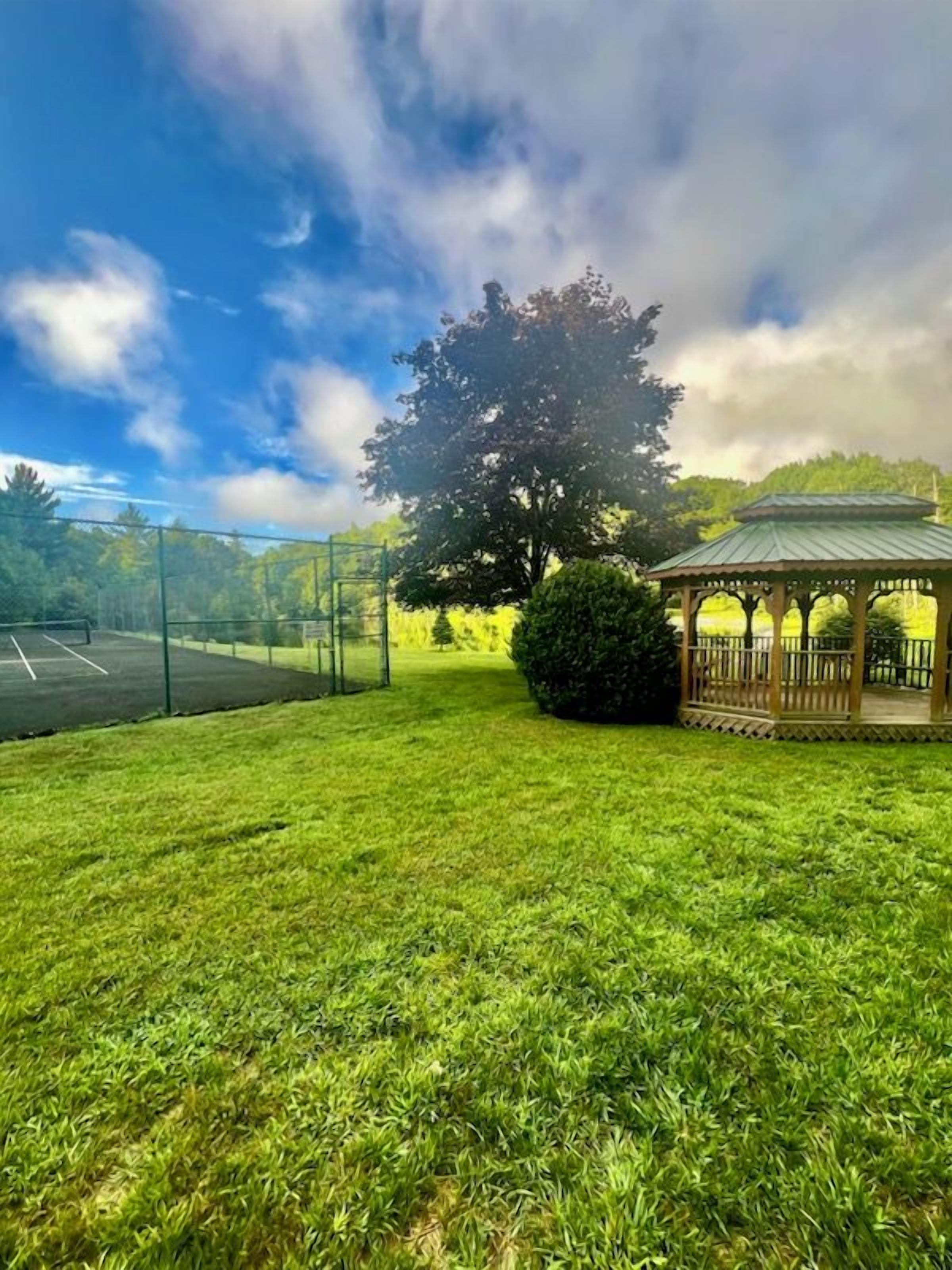 A gazebo with a roof, a fenced tennis court, and a large tree.