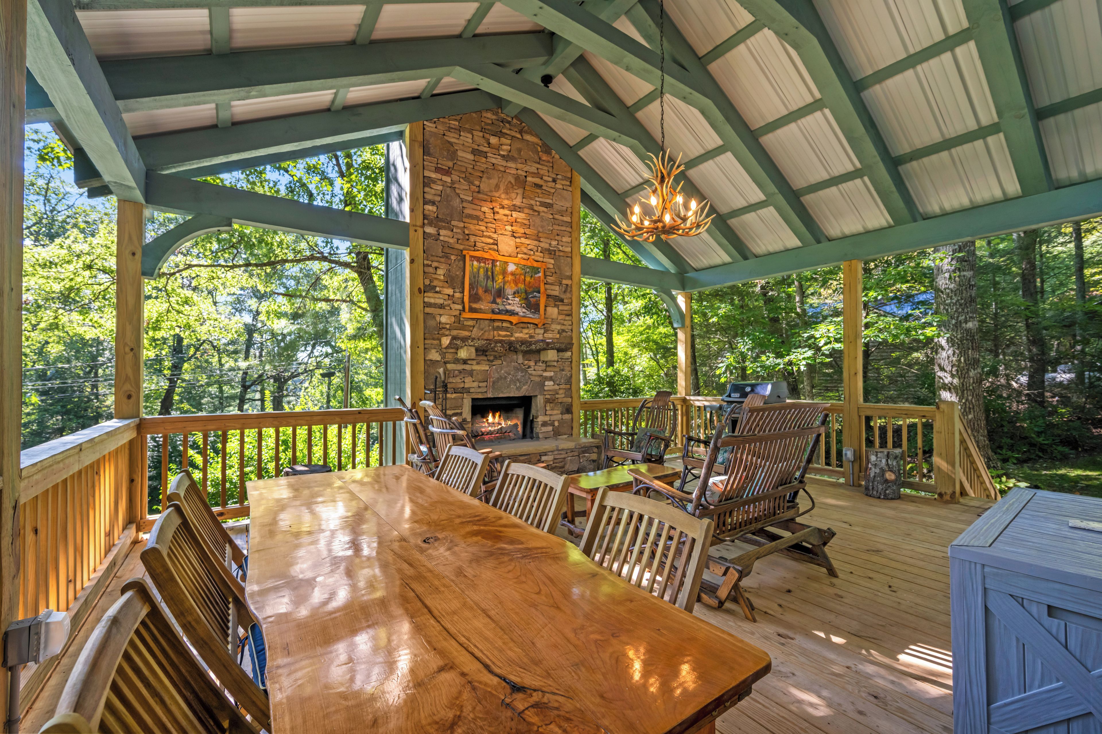 A wooden deck with a stone fireplace, outdoor seating, and a view of the forest.