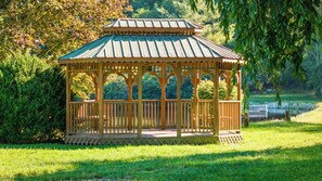 A wooden gazebo with a green roof, surrounded by grass and trees.