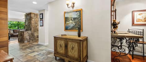 Una habitación amplia con chimenea de piedra, un comedor con mesa y sillas, y un pasillo con un armario de madera y obras de arte.