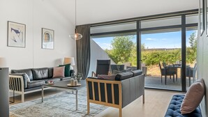 Een moderne woonkamer met een bank, salontafel en een eethoek, met grote ramen en uitzicht op een wijngaard.