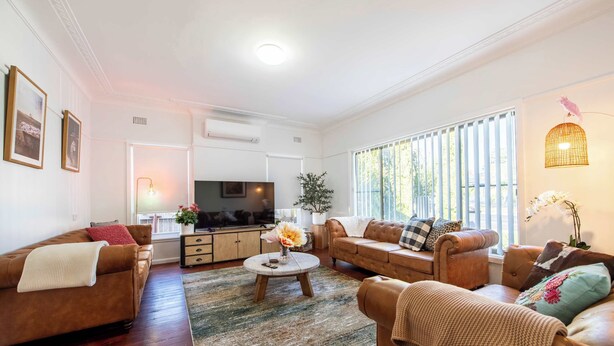 A living room with two sofas, a coffee table, and a TV. There is a window with blinds and a plant. - The Palmer Cove - Stylish, Central (Dubbo)