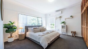 A modern bedroom with a large bed, a bedside table with a lamp, a potted plant, and a window with blinds. - The Palmer Cove - Stylish, Central (Dubbo)