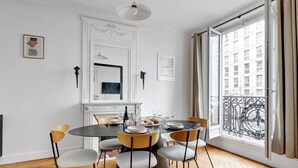 A dining room with a round table, six chairs, a fireplace, and a large window with a view of buildings. - TheBlueStay 318 - Apartment near the Eiffel Tower (Paris)