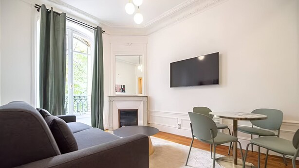 A modern living room with a grey sofa, a fireplace, a flat-screen TV, and a dining area with green chairs.