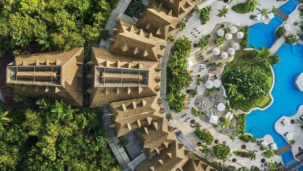 Eine Vogelperspektive zeigt einen luxuriösen Urlaubsort mit einem großen Pool, Palmen und einem markanten Gebäude aus Stein mit einer Wendeltreppe.