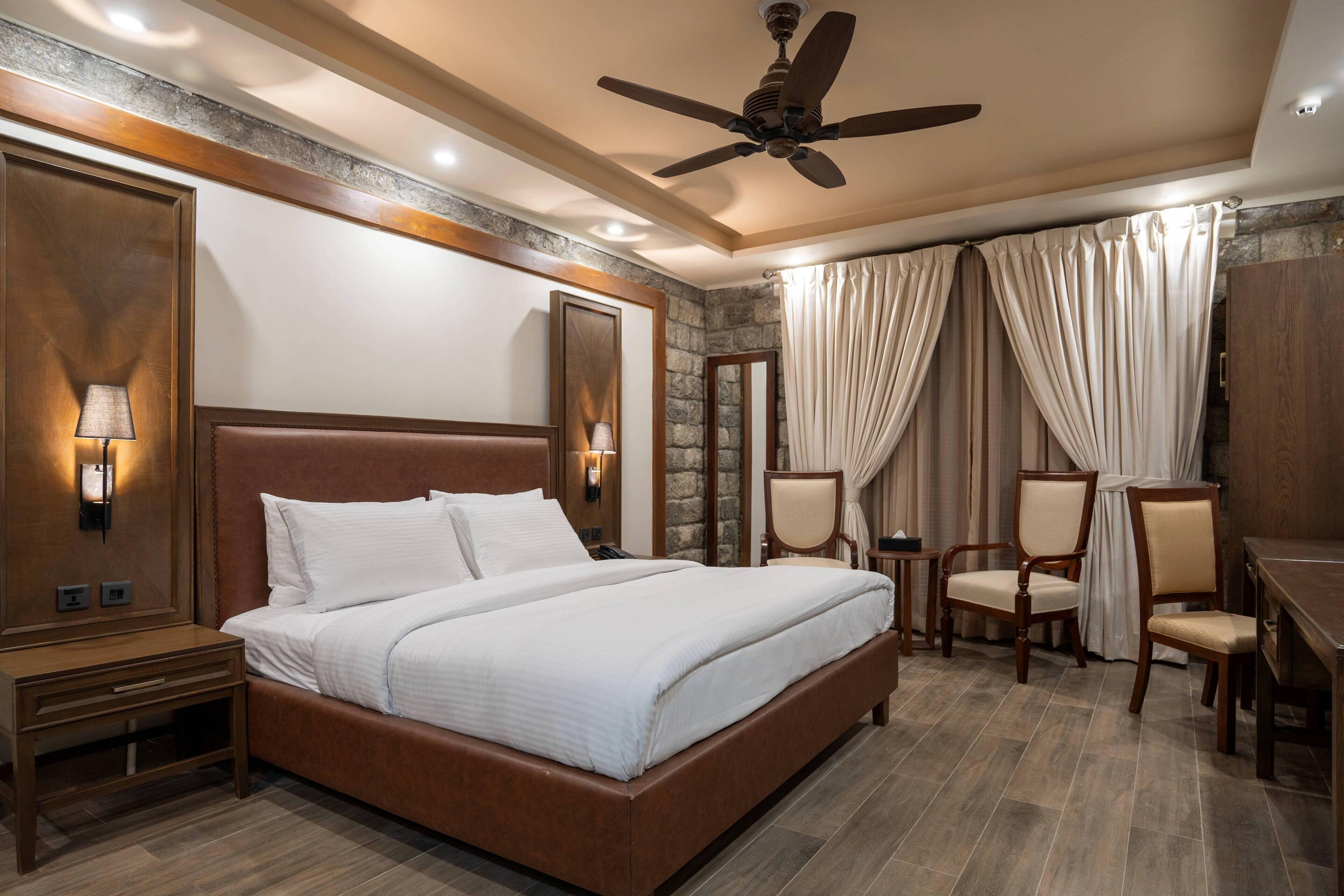 Ein Hotelzimmer mit einem großen Bett, einem Holz-Kopfteil, zwei Nachttischen mit Lampen, einem Schreibtisch und einem Stuhl sowie einem Deckenventilator.
