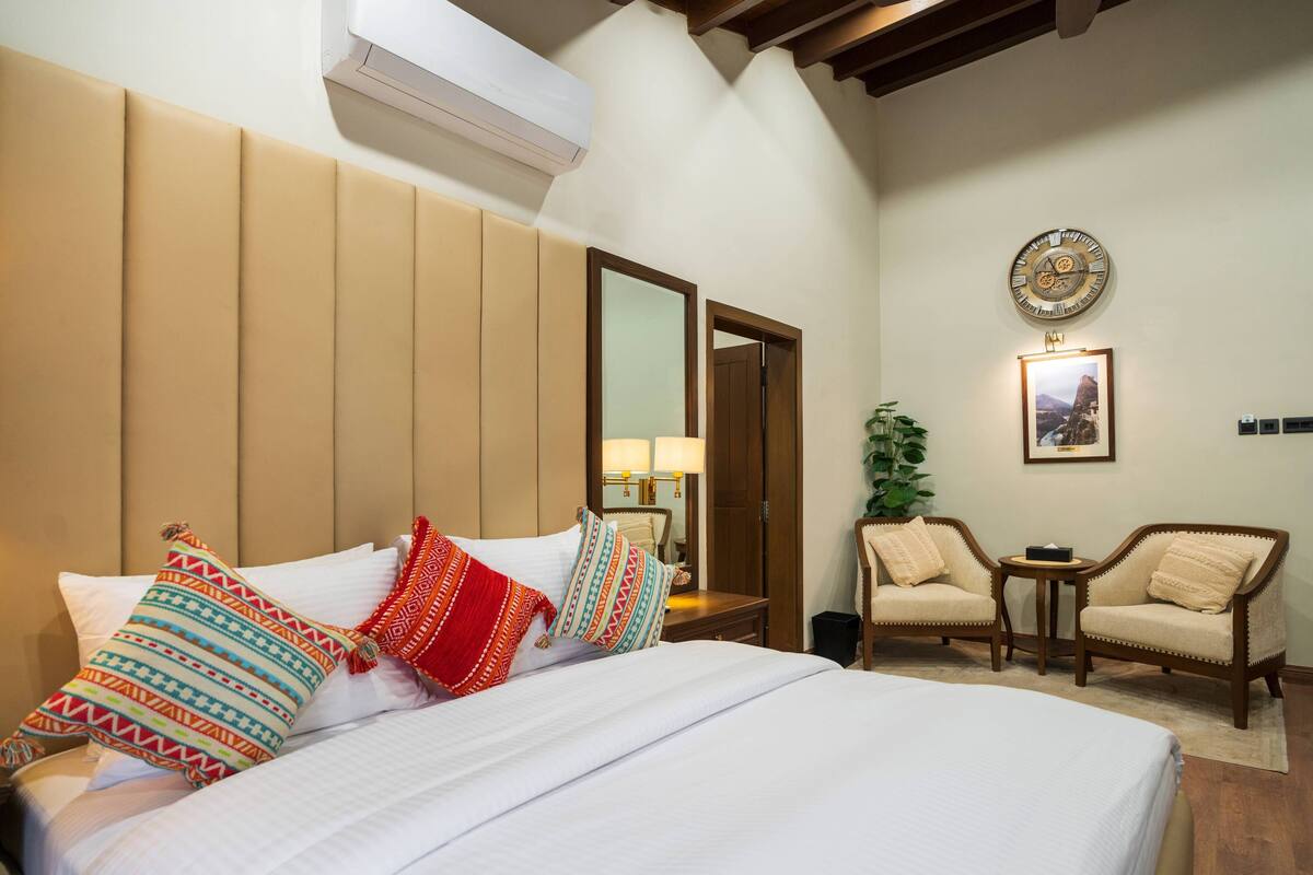 A hotel room with a large bed, a wooden headboard, a wall-mounted air conditioner, a mirror, two armchairs, a small table, and a clock on the wall.