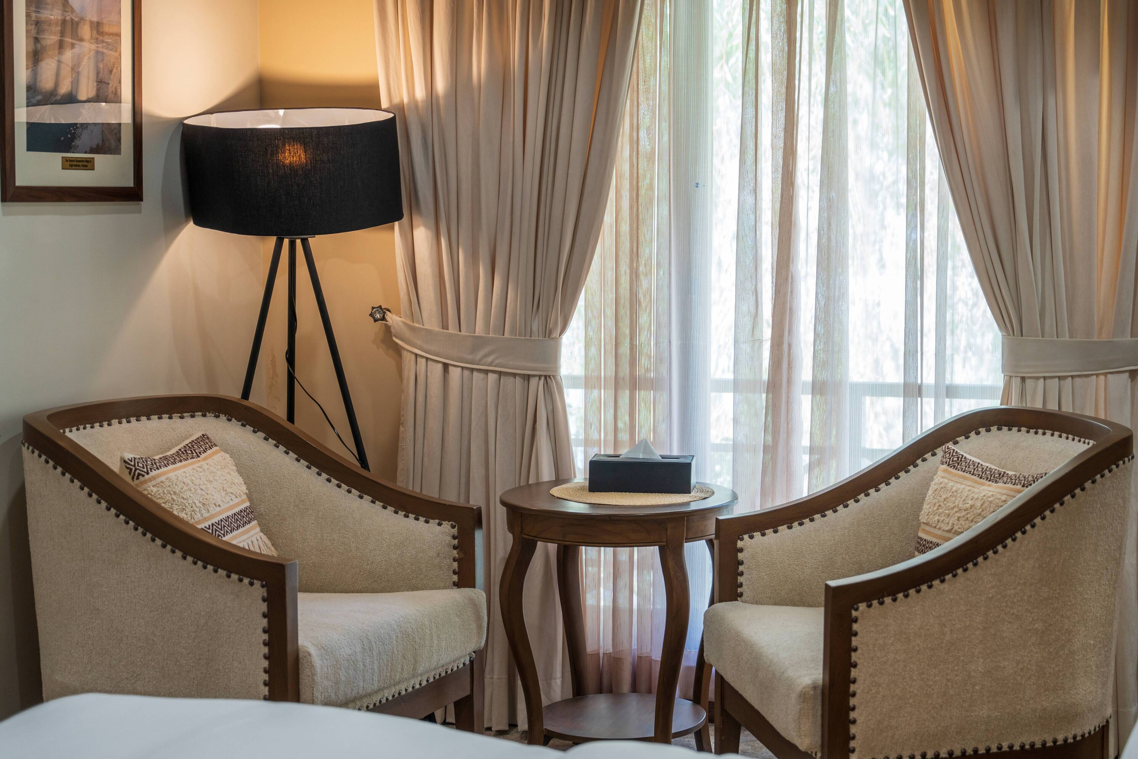 A hotel room with two armchairs, a small round table, a lamp, and a window with curtains.