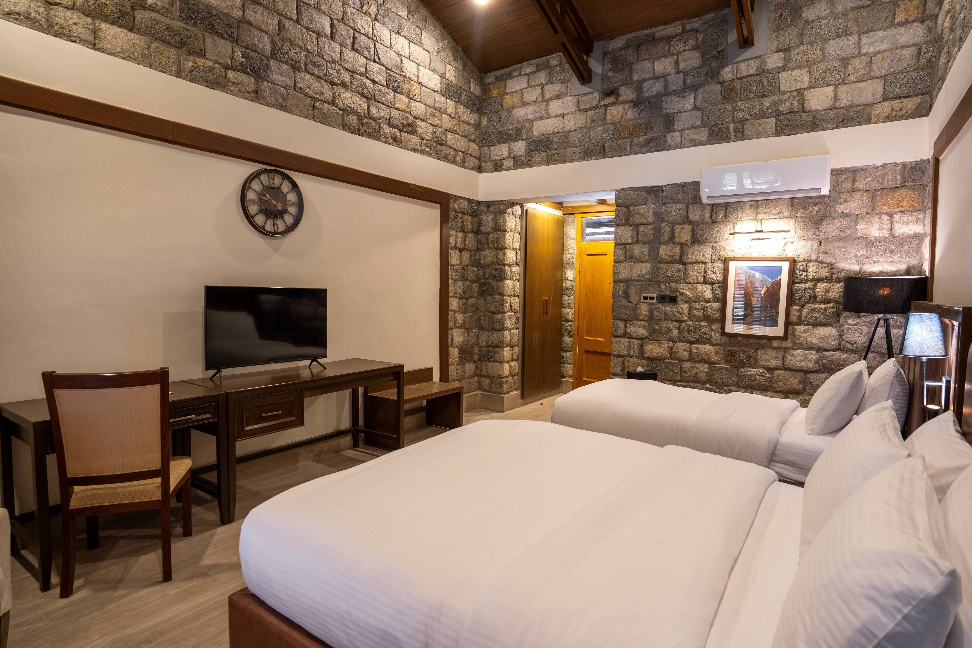 A hotel room with a stone wall, a bed, a desk with a chair, a television, and a door leading to another room.