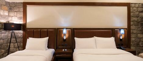 A double room with two beds, wooden headboards, nightstands, lamps, and a stone wall.