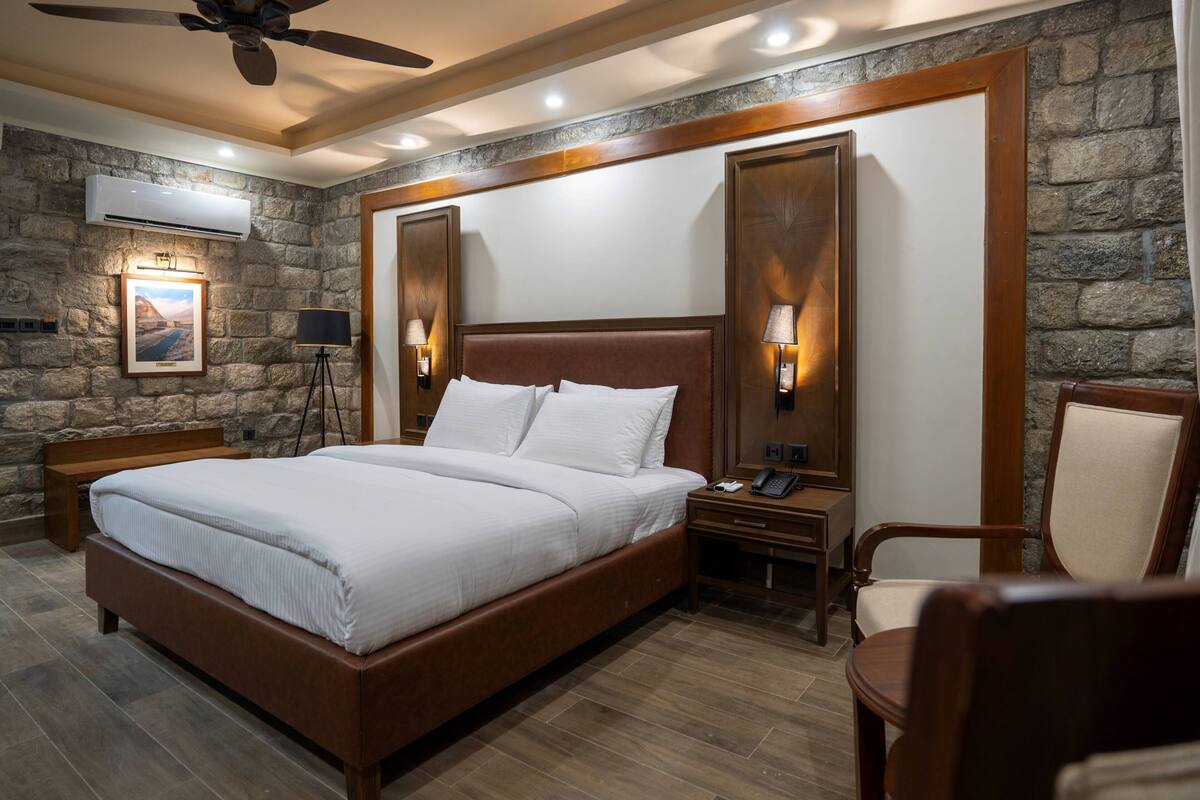 A hotel room with a large bed, wooden headboard, stone wall, and a ceiling fan.
