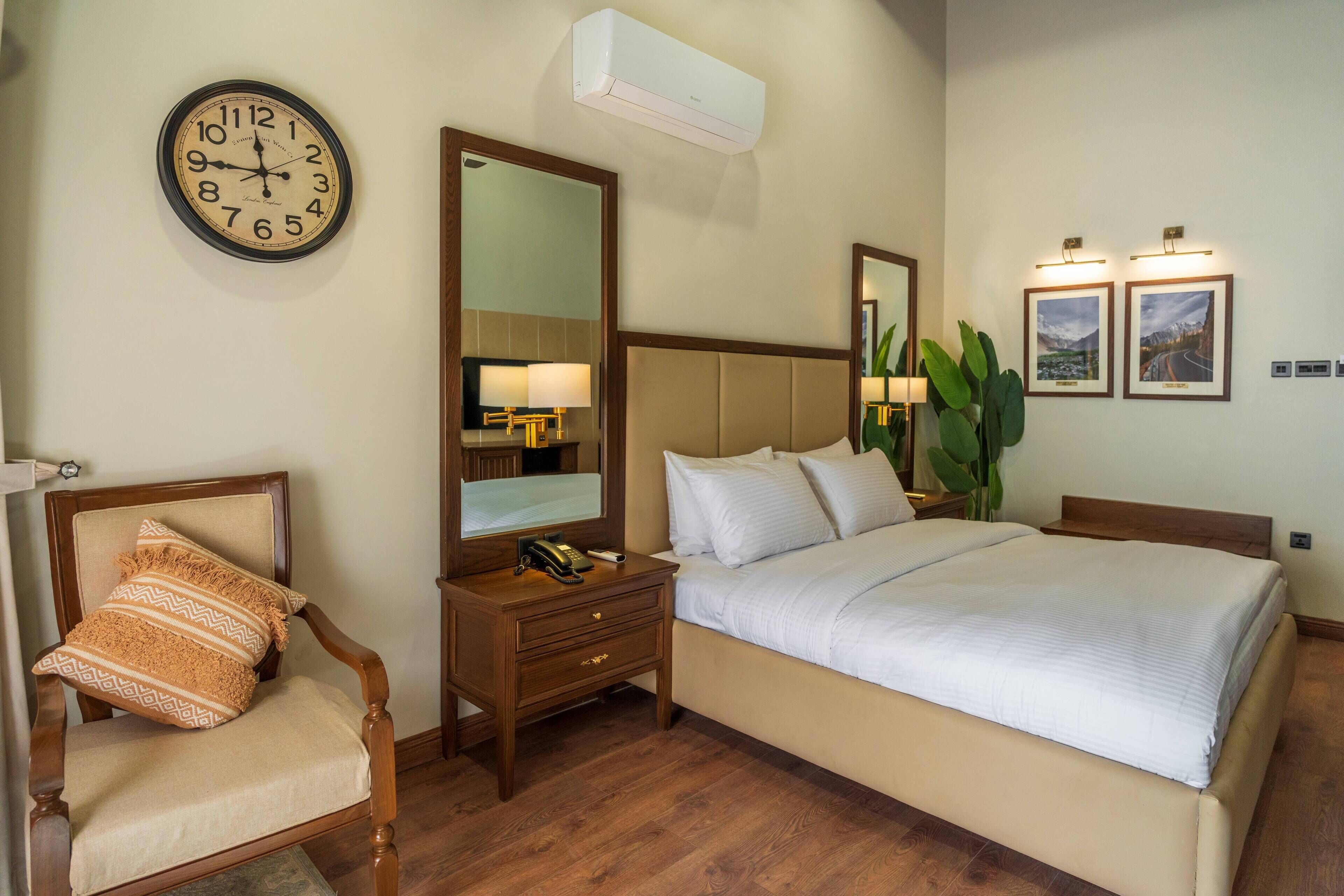 A hotel room with a bed, a chair, a nightstand, a clock, a mirror, and two framed pictures on the wall.