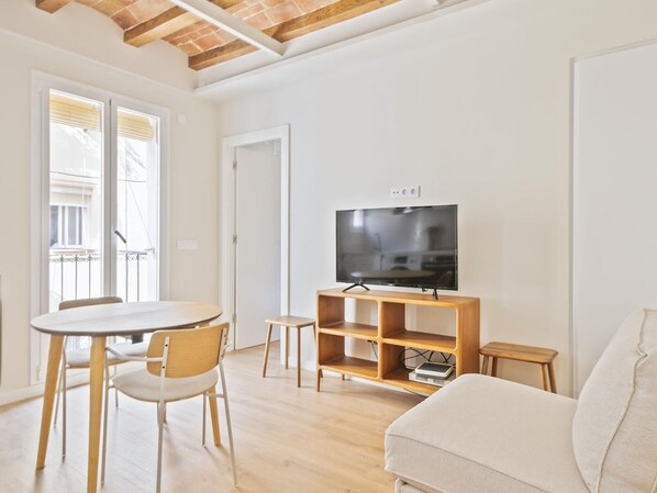 Un salon avec un téléviseur posé sur un meuble en bois, un canapé, une table à manger avec des chaises et un balcon doté d’une balustrade.