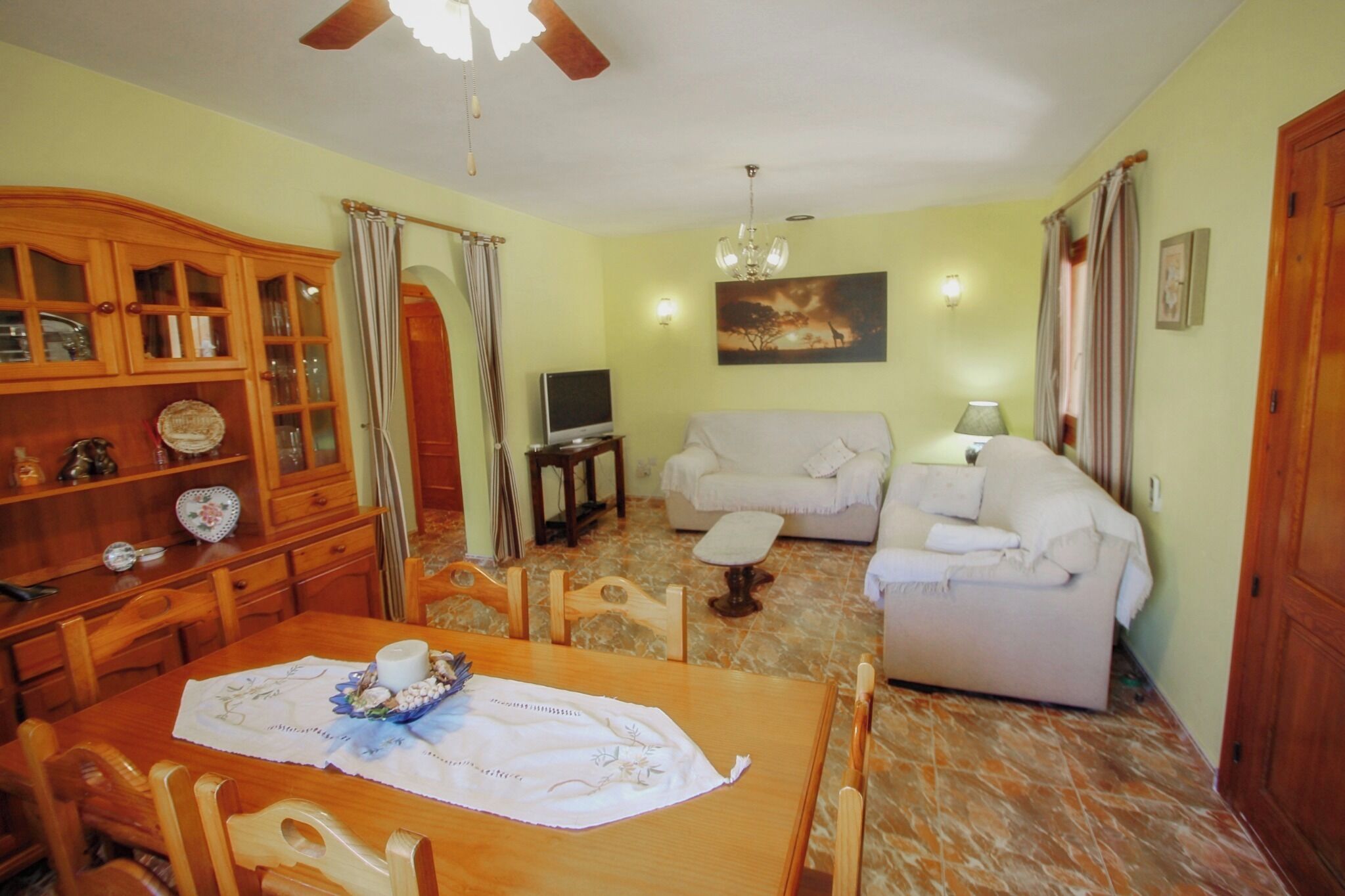 A living room with a sofa, a TV, a dining table with chairs, a wooden cabinet, and a ceiling fan.