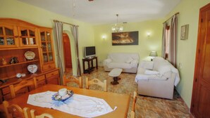 A living room with a sofa, a TV, a dining table with chairs, a wooden cabinet, and a ceiling fan. - Finca Pineda - private pool villa in Benissa (Benissa)