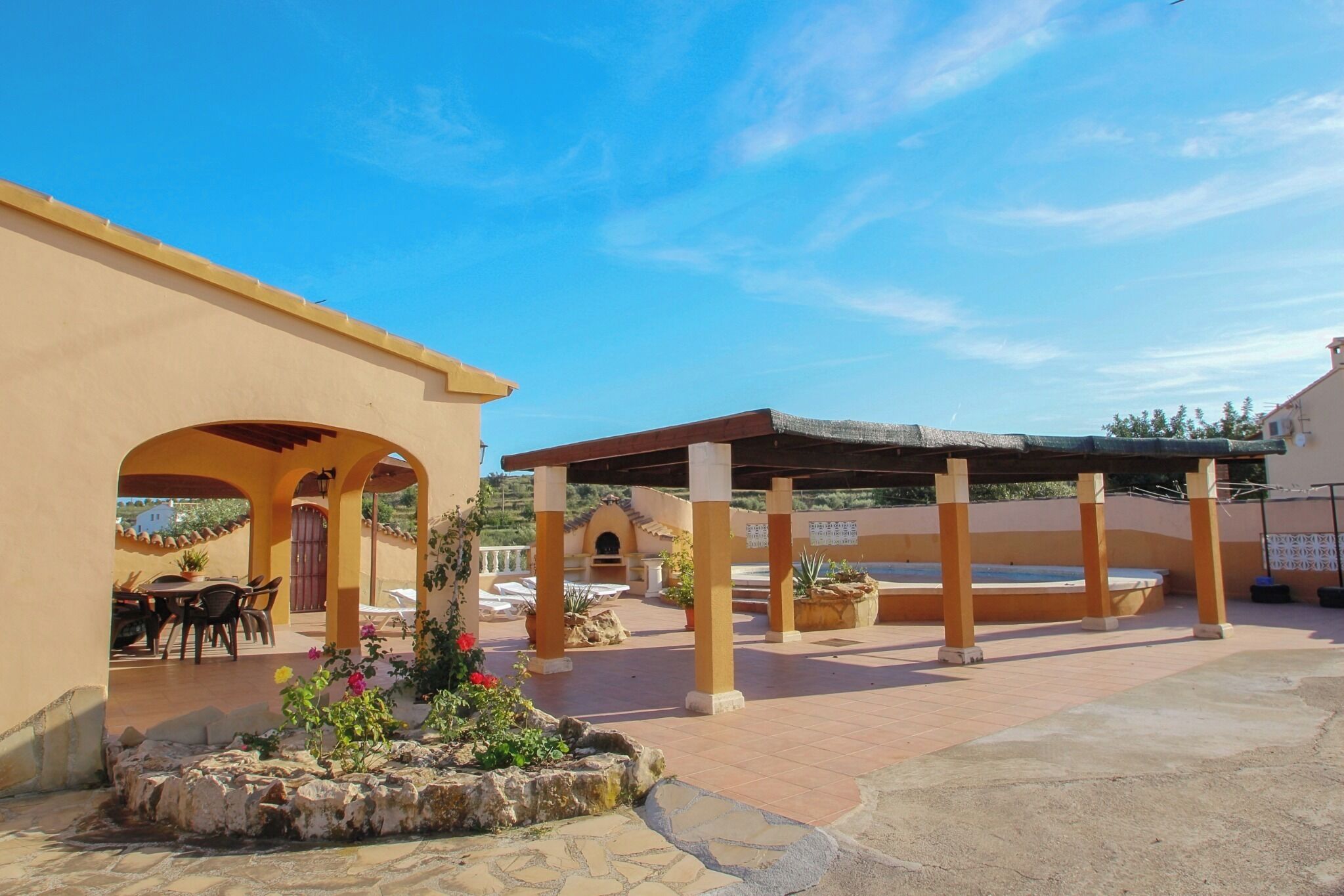 An outdoor patio area with a covered seating area, stone planters with flowers, and a pool.