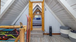 A cozy attic bedroom with a sloped ceiling, a bed with blue bedding, and a wooden ladder leading to a lofted sleeping area. - Tee & Ski Cabin | Mt Rainier, White Pass (Packwood)