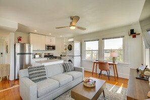 En moderne stue med en grå sofa, et sofabord og et spiseområde med en stol og et bord. Et køkken med hårde hvidevarer og en ventilator er synligt i baggrunden.